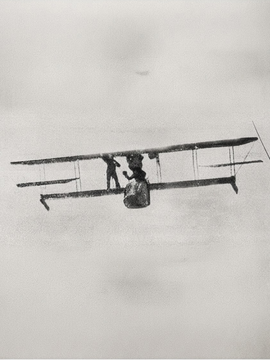 A biplane in flight against a pale sky, a man standing on its lower wing beside the pilot