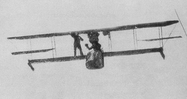 A biplane in flight against a pale sky, a man standing on its lower wing